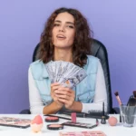 pleased-pretty-caucasian-woman-sitting-table-with-makeup-tools-holding-money